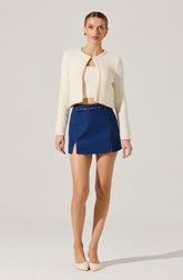 A woman is wearing a cream-colored, textured, long-sleeve, cropped cardigan with a round neckline and a small opening at the front. Underneath, she has a matching knit top. She pairs this with a high-waisted, dark blue mini skirt featuring two front slits and a small belt at the waist. She completes the outfit with light-colored pointed-toe heels.